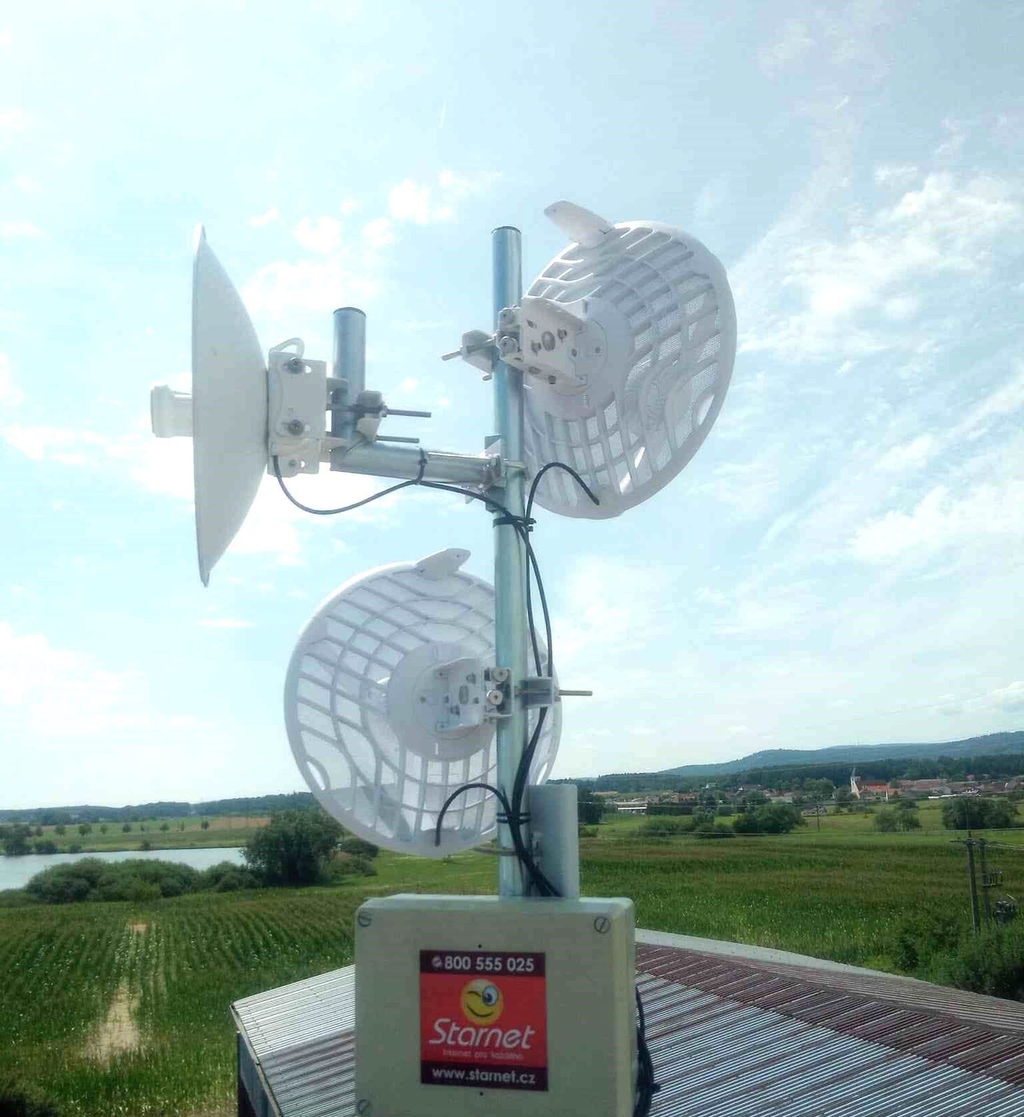 Mřížkové parabolické antény na střeše, v pozadí pole a krajina